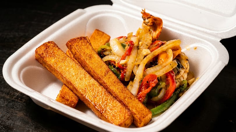 Takeout container with golden-brown fries and sautéed vegetables, including onions, peppers, and mushrooms.