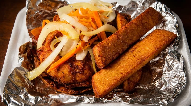 Fried chicken, yuca fries, and pickled onions with carrots in a foil-lined takeout container.