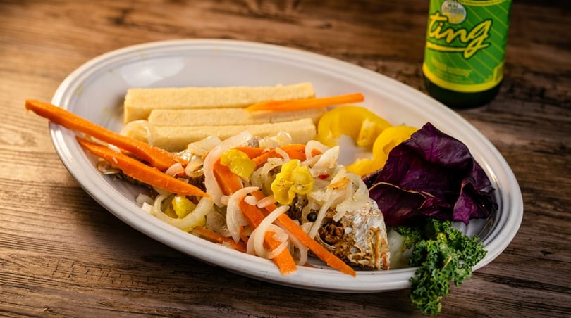 Escovitch fish with pickled vegetables, bammy, and a Ting soda on a wooden table.
