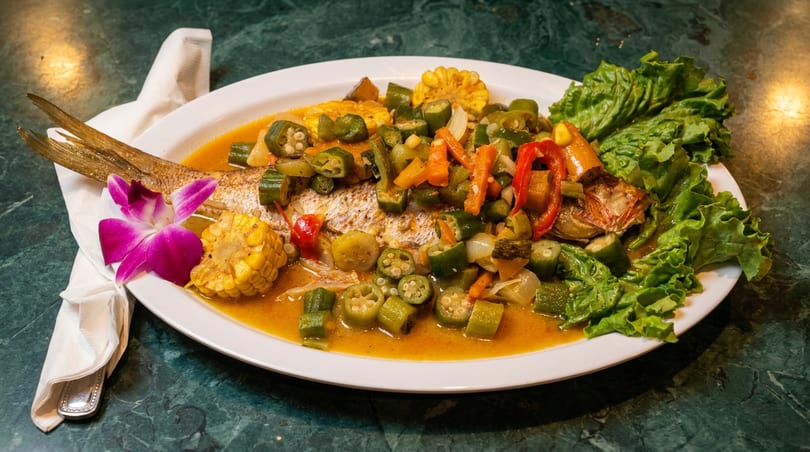 Whole fish dish with okra, corn, peppers, and lettuce on a white plate with a purple orchid.